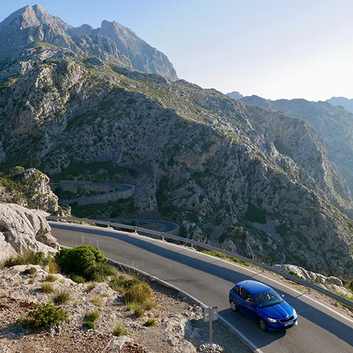 Coche azul en carretera de monta&ntilde;a con paisaje rocoso y monta&ntilde;as al fondo.