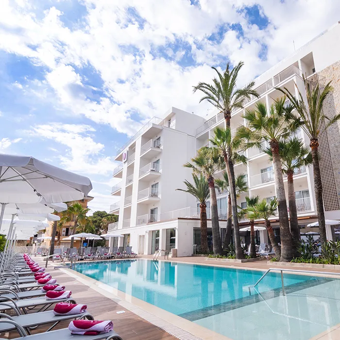 Piscina junto a un hotel con palmeras y cielo parcialmente nublado.