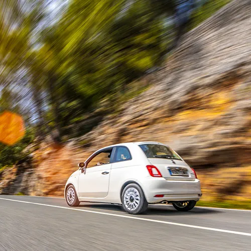 Auto blanco compacto circulando por una carretera junto a un muro de piedra.