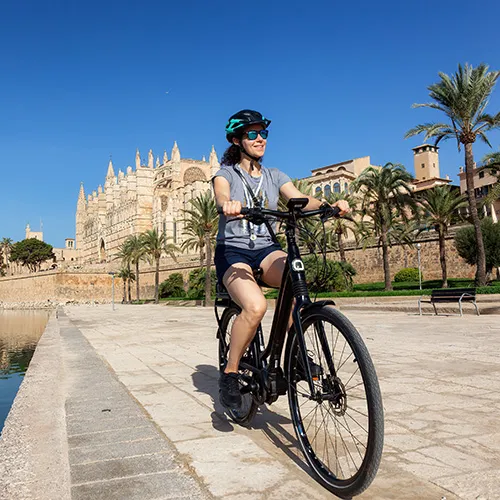 Persona andando en bicicleta cerca de una catedral y palmeras bajo un cielo azul.