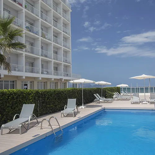 Piscina al lado de un hotel con tumbonas y sombrillas bajo un cielo azul.