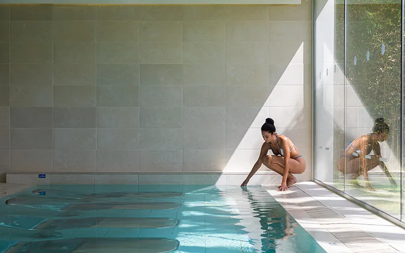Mujer tocando el agua de una piscina interior iluminada por el sol.