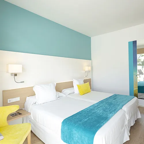 Habitaci&oacute;n moderna con dos camas, acentos turquesas y silla amarilla.