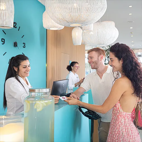 Personas en recepci&oacute;n de hotel saludan y sonr&iacute;en frente a un mostrador azul.