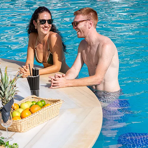 Pareja conversando en la piscina junto a una canasta de frutas.