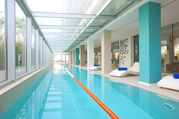 Piscina interior con tumbonas blancas y techo de vidrio.