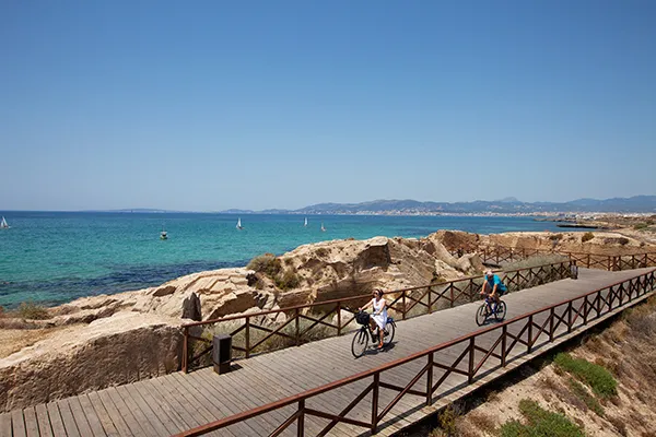 Dos personas en bicicleta por un paseo junto al mar en un d&iacute;a soleado.