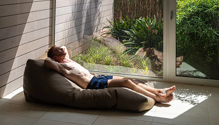 Hombre recostado en un puff, disfrutando del sol junto a una puerta de vidrio.