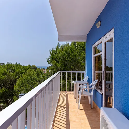 Balc&oacute;n con sillas blancas y vista a &aacute;rboles, fachada azul, cielo despejado.
