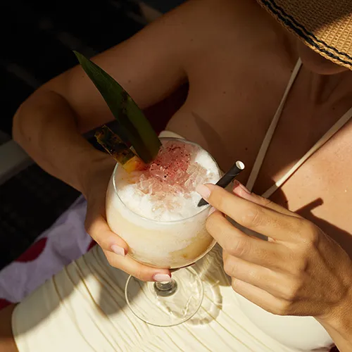 Persona disfrutando un c&oacute;ctel tropical con sombrero y traje de ba&ntilde;o claro.