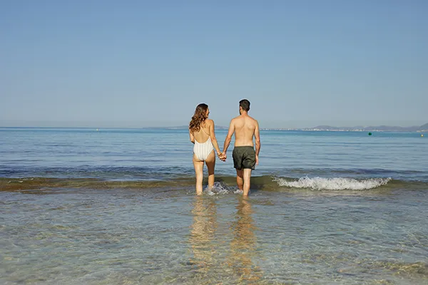 Pareja caminando hacia el mar, agarrados de la mano, en una playa soleada.