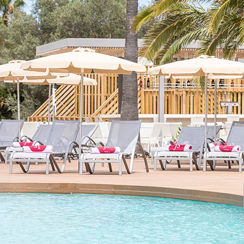 Sombrillas y tumbonas junto a la piscina en un entorno de palmeras.