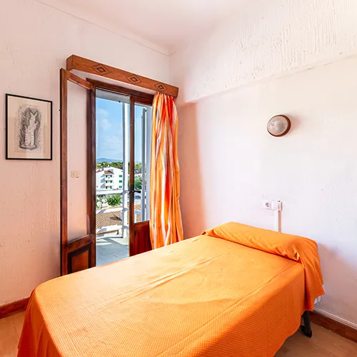 Habitaci&oacute;n minimalista con cama anaranjada y vista a la ciudad desde la ventana.