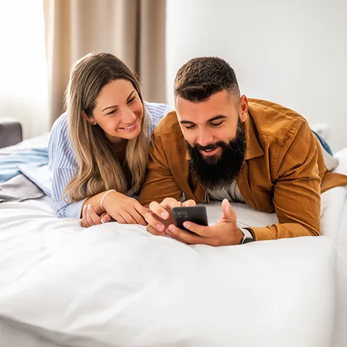 Pareja sonr&iacute;e mientras mira un celular recostada en una cama.