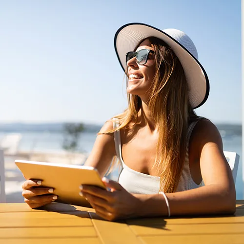Mujer con sombrero y gafas de sol sonr&iacute;e al aire libre con una tableta.