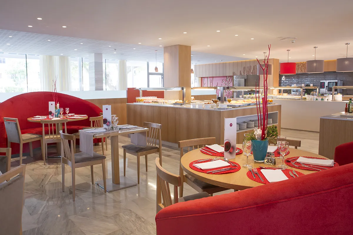 Restaurante luminoso con mesas rojas y buffet en el fondo.