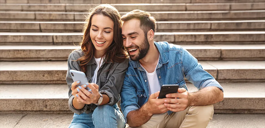 Pareja sonriente mirando sus tel&eacute;fonos en unas escaleras.