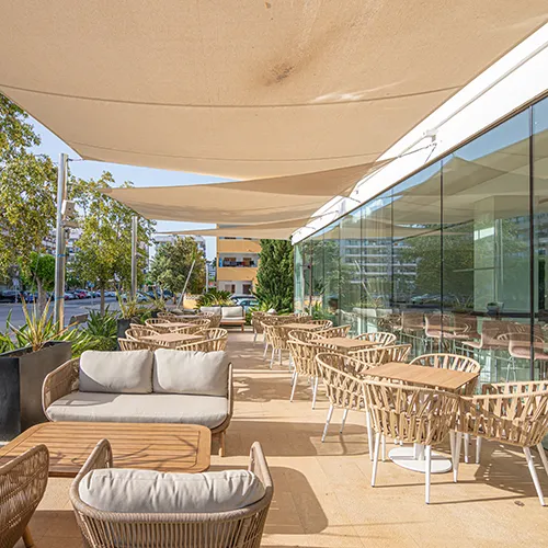 Terraza con mesas, sillas tejidas y sof&aacute;s bajo toldos, junto a un edificio de vidrio.