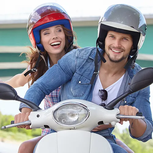 Pareja sonriente con cascos monta una scooter blanca frente a un puente.