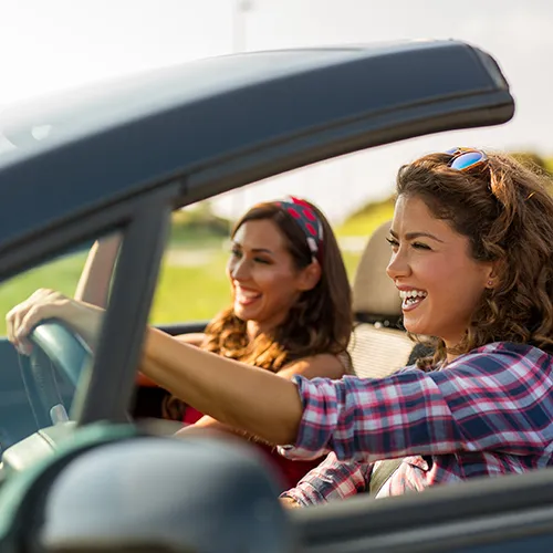 Dos mujeres sonr&iacute;en mientras conducen un coche descapotable bajo el sol.