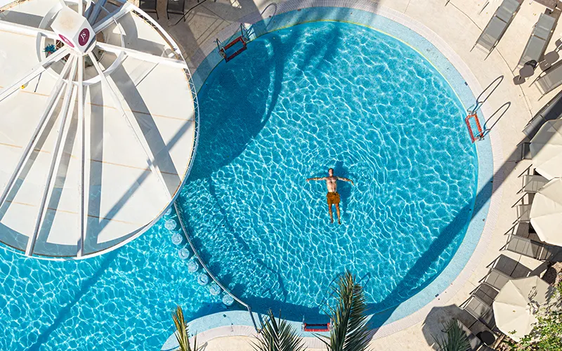Hombre flotando en piscina redonda, vista a&eacute;rea, sol y sombras alrededor.