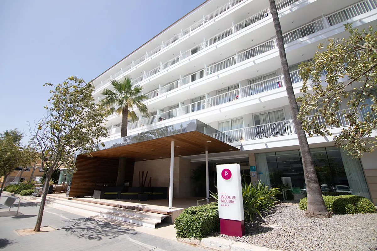 Fachada de hotel moderno con balcones y vegetaci&oacute;n en la entrada.