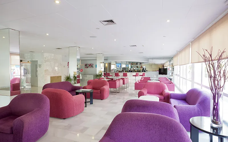 Sala de estar moderna con sof&aacute;s morados y mesas cerca de ventanas grandes.