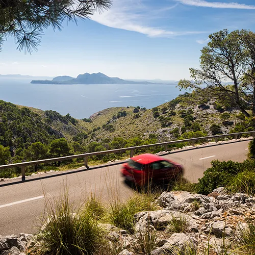 Coche rojo en carretera costera con paisaje monta&ntilde;oso y mar al fondo.