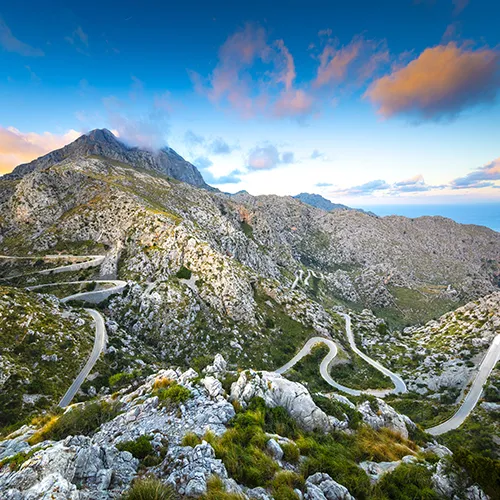Camino serpenteante en un paisaje monta&ntilde;oso bajo un cielo azul y nubes rosadas.