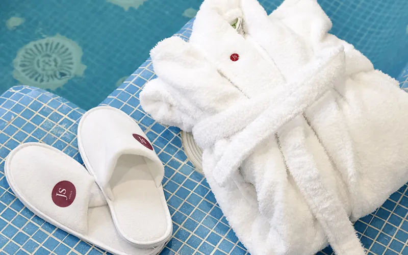 Bata y pantuflas blancas sobre una superficie de azulejos azules junto a una piscina.