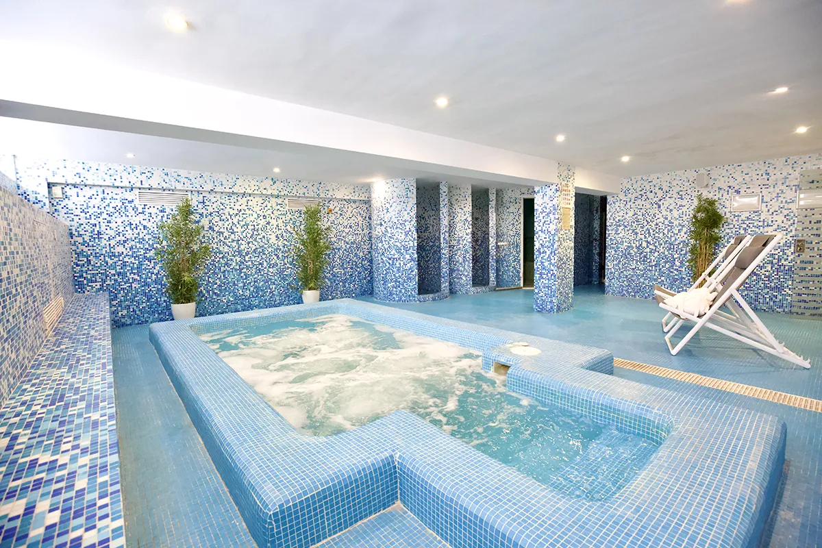 Spa con jacuzzi azul y tumbona, adornado con azulejos y plantas.