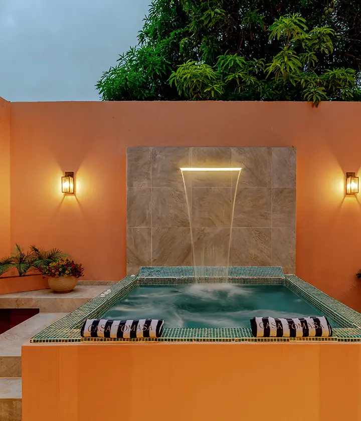 Jacuzzi en patio con cascada, toallas a rayas y luz tenue, rodeado de vegetación.