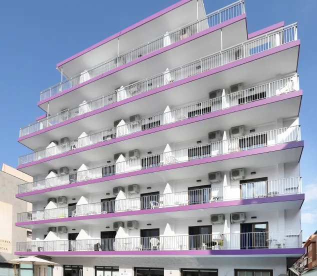 Edificio de seis pisos con balcones y detalles morados, frente a un cielo despejado.