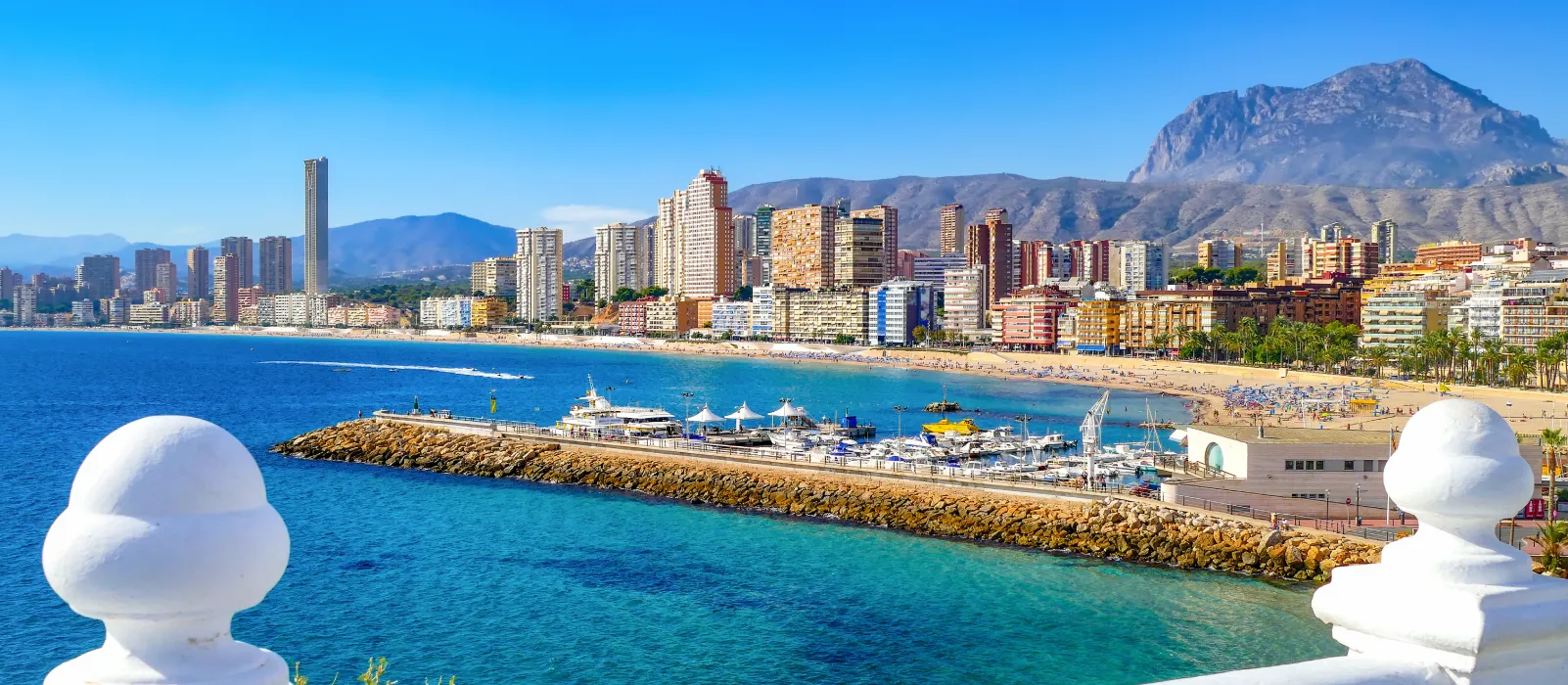 Vista panorámica de la costa con edificios, playa y montañas al fondo.