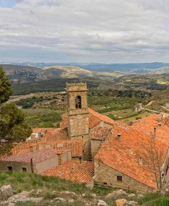 Tejado rojo en un pueblo con campanario y paisaje montañoso al fondo.