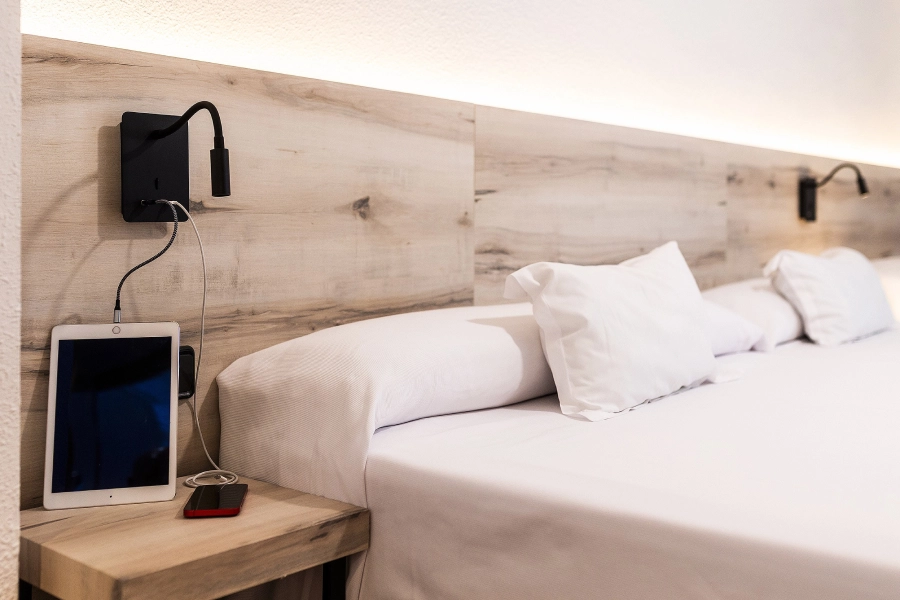 Cama moderna con tablets y lámparas en la cabecera de madera clara.