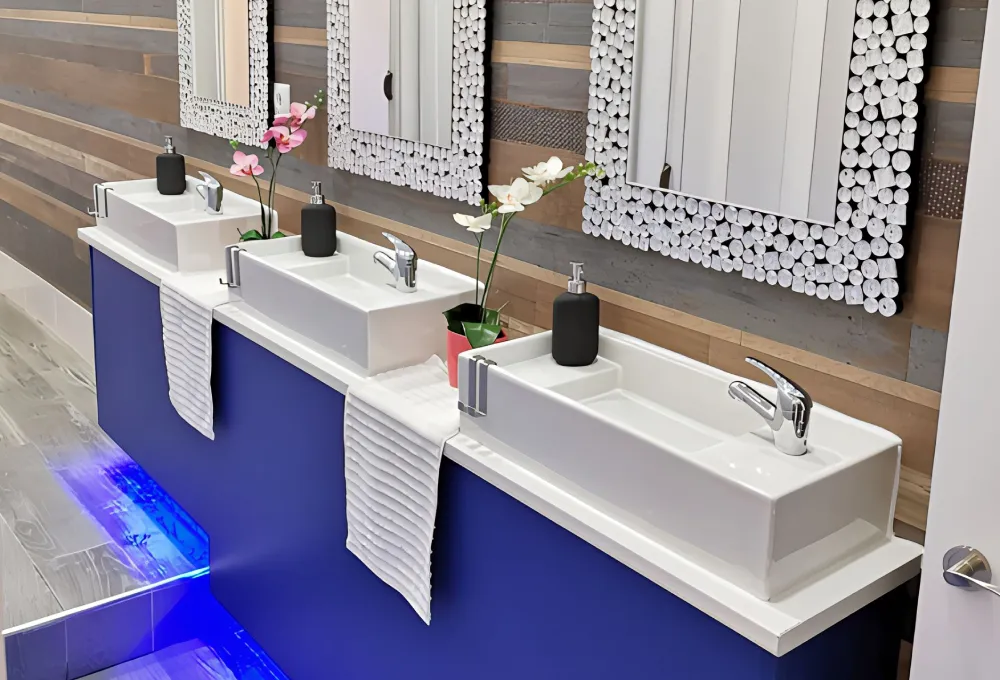 Tres lavabos modernos con espejos decorativos, dispensadores y flores sobre encimera azul.