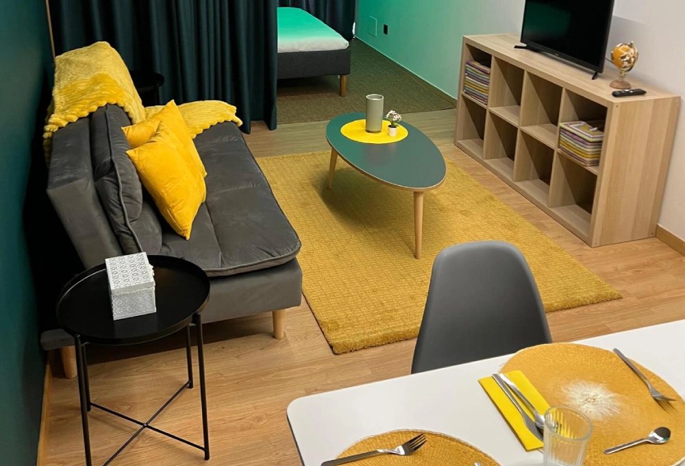 Sala moderna con sof&aacute; gris, cojines amarillos, mesa de centro, tapete amarillo y estanter&iacute;a.