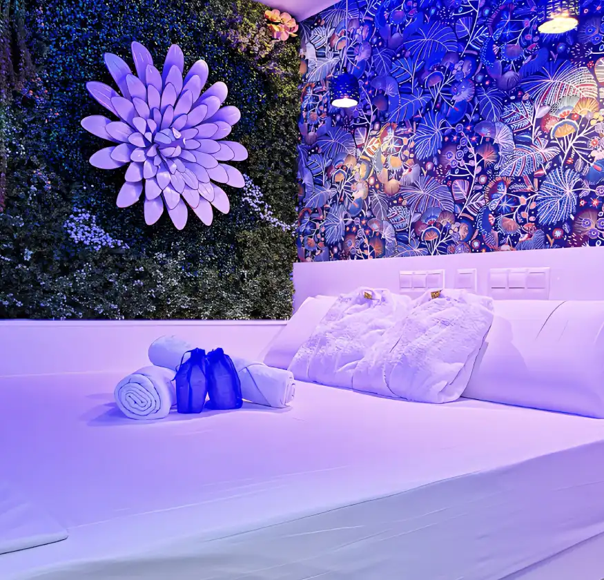 Cama con s&aacute;banas blancas, toallas enrolladas y un mural floral iluminado en tonos azules.