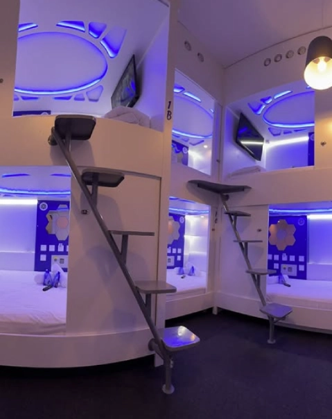 C&aacute;psulas futuristas iluminadas con luces azules y escaleras en un albergue moderno.
