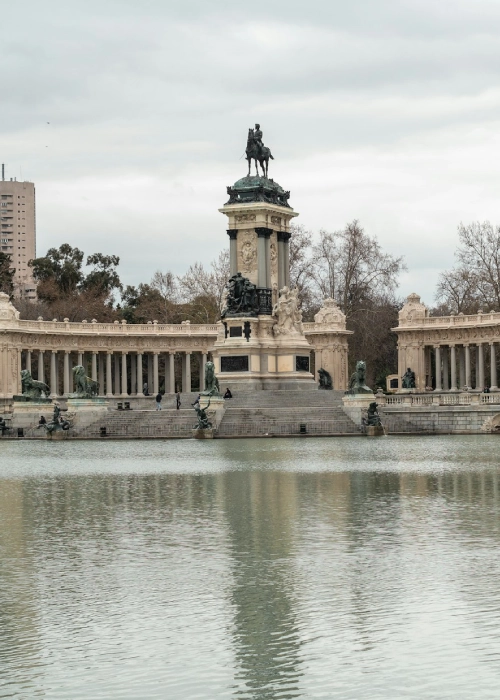 Estanque y monumento en un parque urbano, con &aacute;rboles y cielos nublados al fondo.