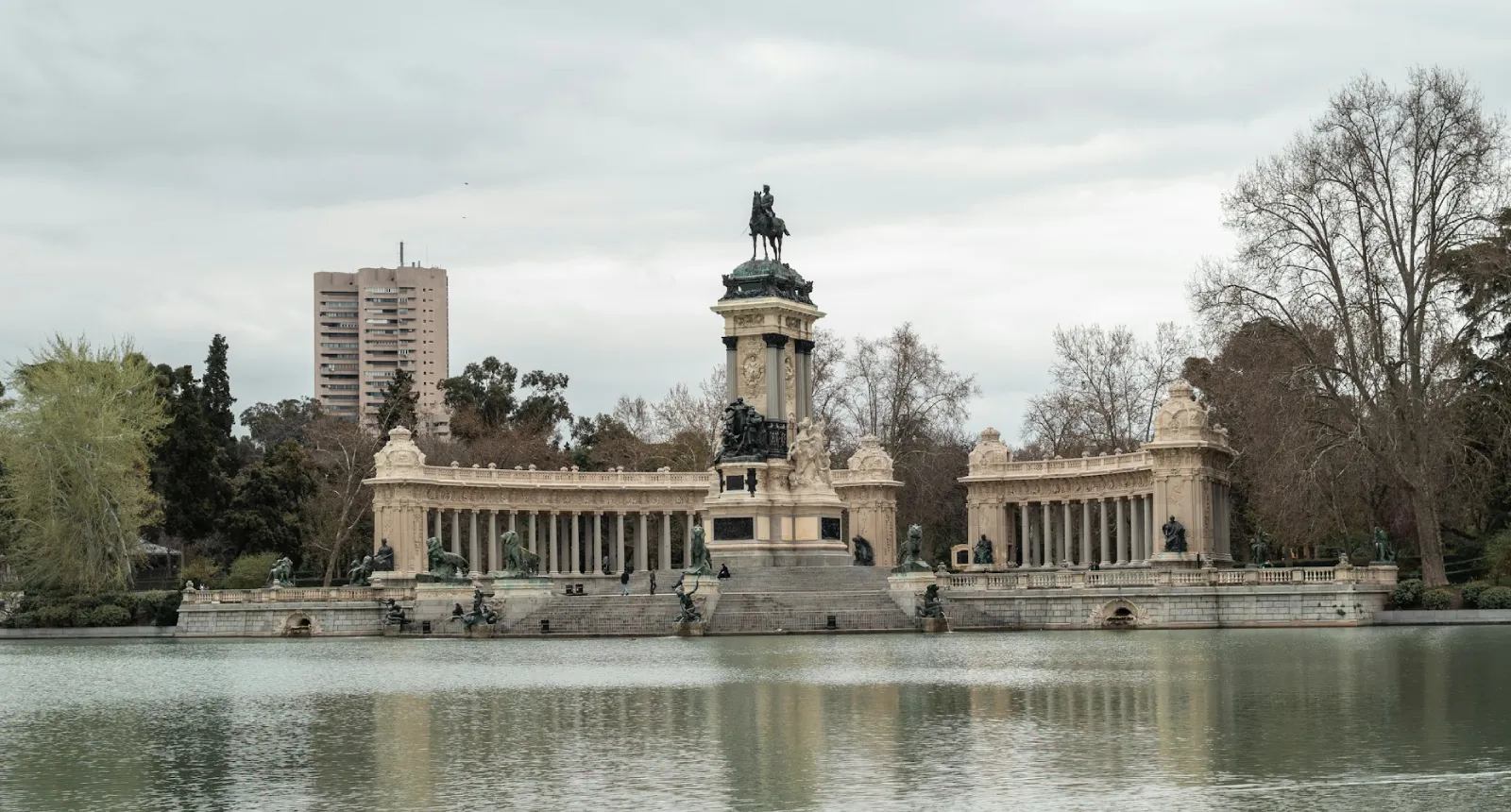Monumento junto a un lago con &aacute;rboles y un edificio alto al fondo bajo cielo nublado.