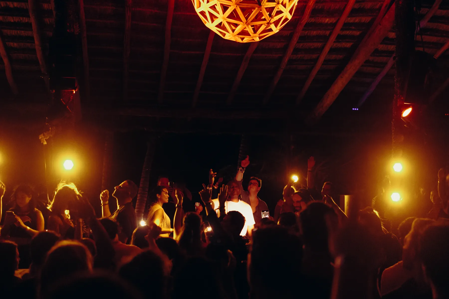 Personas bailando en una fiesta con luces cálidas y techo de madera.