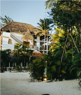 Casa tropical con techo de paja rodeada de palmeras y arena blanca.