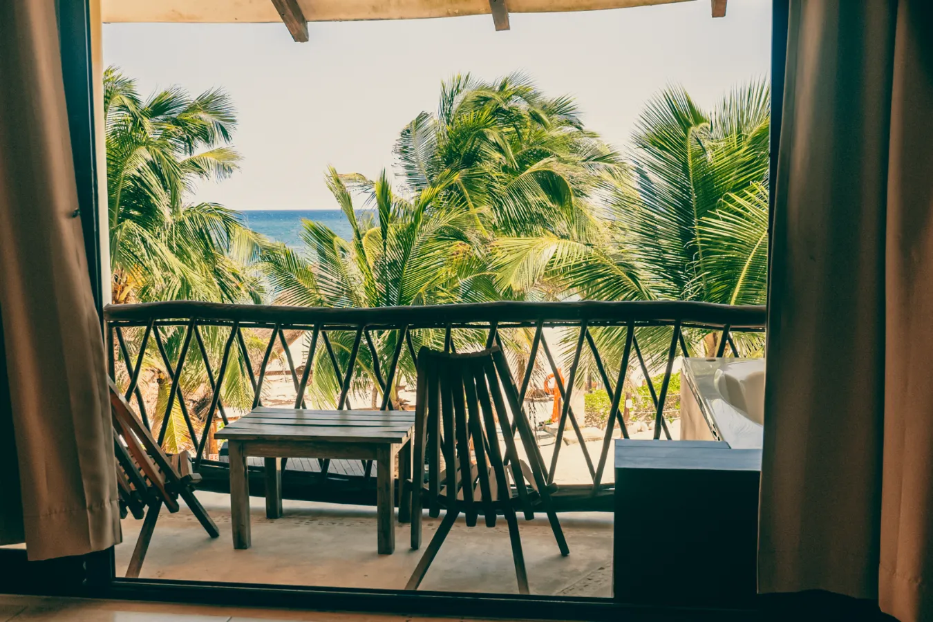Balcón con sillas, mesa y vista al mar entre palmeras.