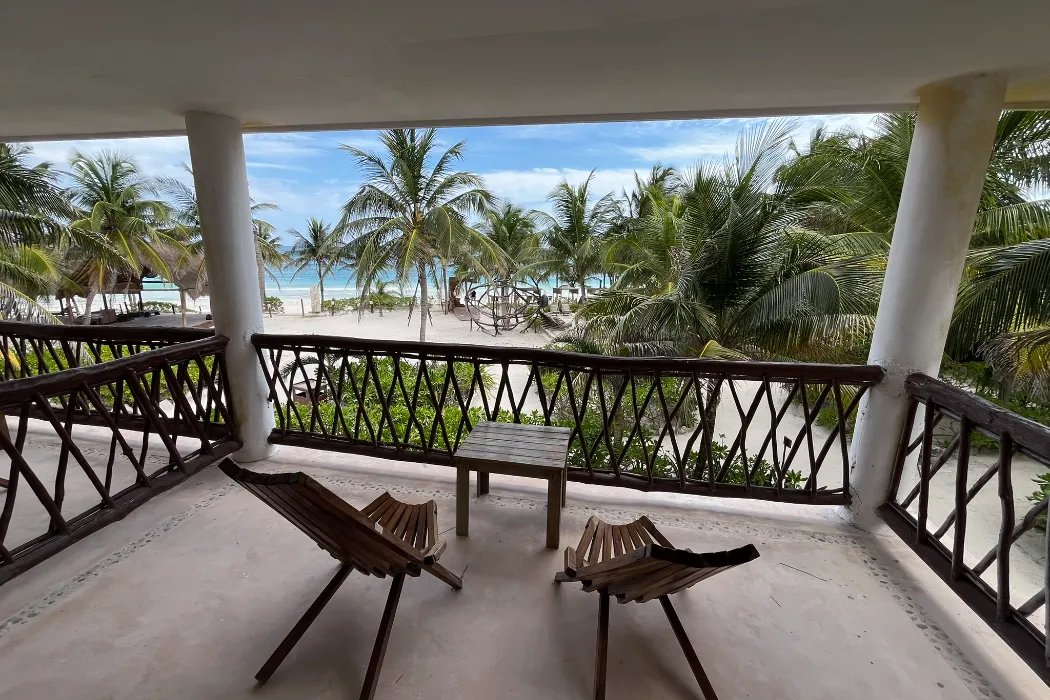 Balcón con dos sillas, mesa y vista a la playa con palmeras y el mar.