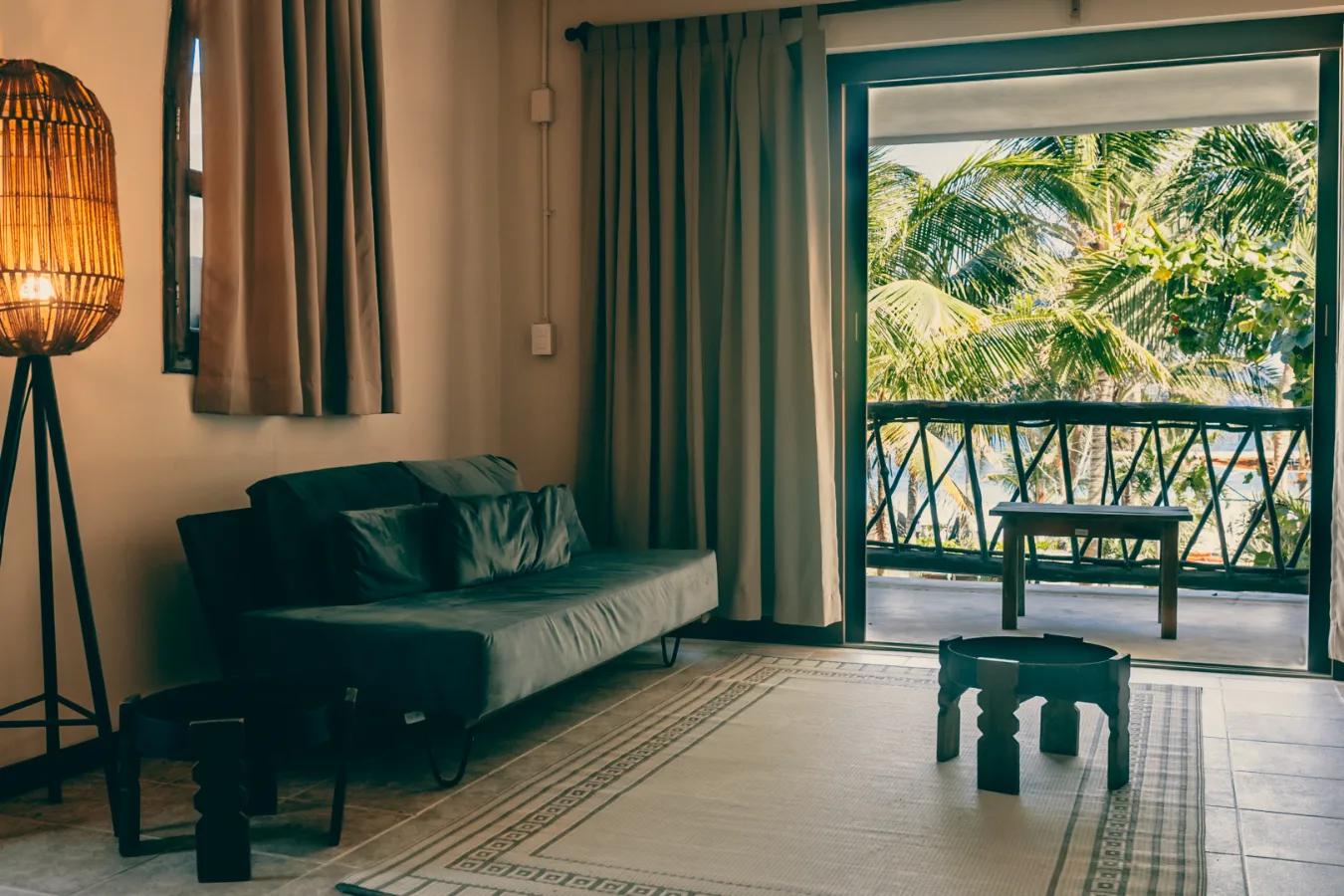 Sala con sof&aacute;, l&aacute;mpara y balc&oacute;n hacia palmeras, iluminaci&oacute;n c&aacute;lida y acogedora.
