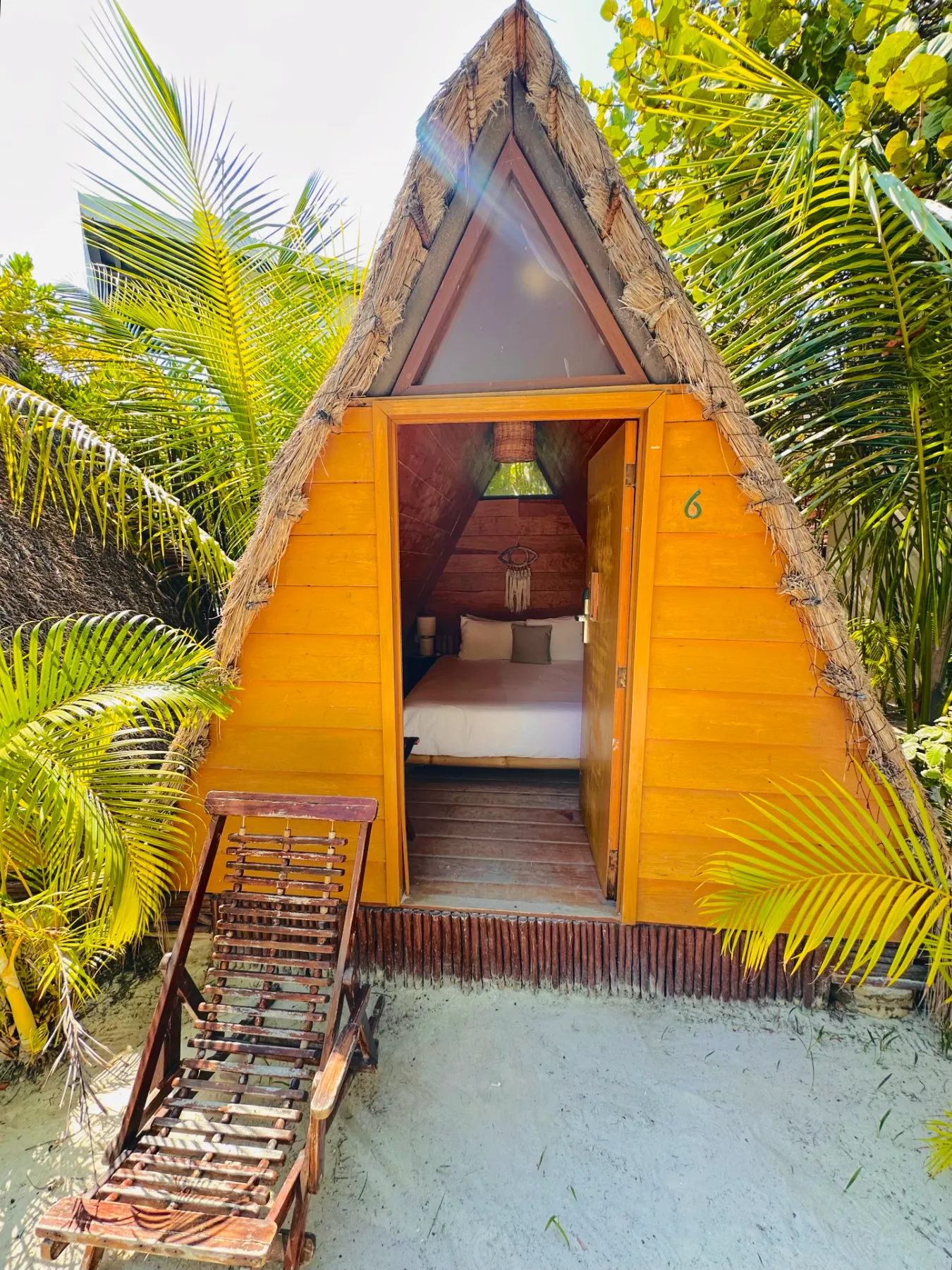 Cabaña triangular de madera entre palmeras con una tumbona.