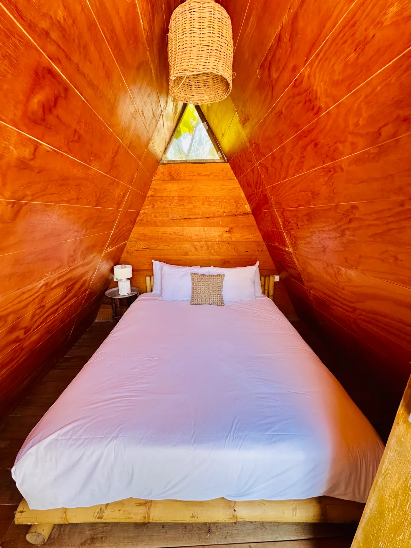 Caba&ntilde;a triangular de madera con cama y l&aacute;mpara de mimbre.