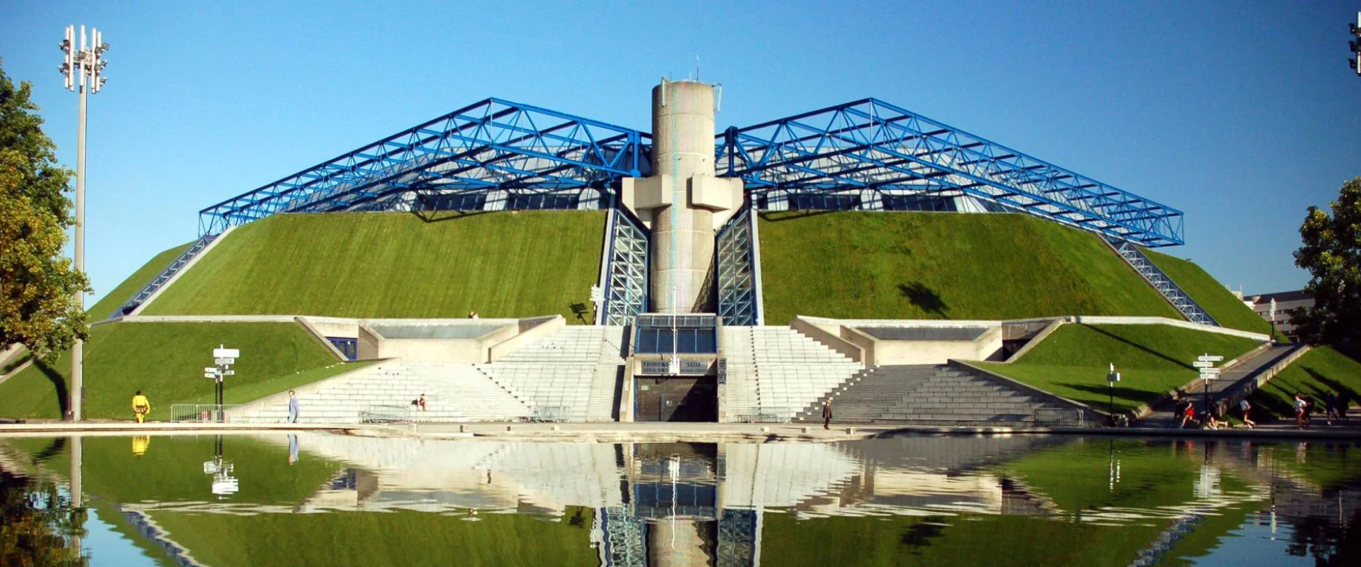 Bâtiment moderne avec structure bleue et toit vert, reflet dans l'eau.